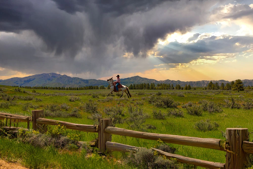 A person riding a horse along a wooden fence line under dramatic Montana storm skies
